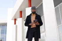 Bajo ángulo positivo afroamericano masculino en elegante desgaste y máscara de la cara tableta de navegación mientras está de pie en la calle soleada - foto de stock