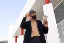 Low angle positive African American male in stylish wear and face mask doing video call on tablet while with clenched fist celebrating victory with gesture standing on sunny street — Stock Photo