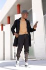 Cheerful African American male in stylish clothes and earbuds browsing modern tablet on sunny street and looking at screen with smile — Stock Photo