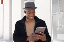 Alegre hombre afroamericano en ropa elegante y auriculares navegando tableta moderna en la calle soleada y mirando a la pantalla con sonrisa - foto de stock