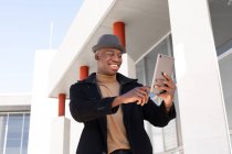 Alegre hombre afroamericano en ropa elegante y auriculares navegando tableta moderna en la calle soleada y mirando a la pantalla con sonrisa - foto de stock