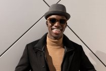 Moda confiante feliz afro-americano masculino em chapéu e óculos de sol em pé contra a parede cinza e olhando para a câmera — Fotografia de Stock