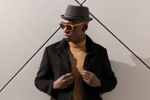 Hombre afroamericano confiado de moda en sombrero y gafas de sol de pie contra la pared gris y mirando hacia otro lado - foto de stock