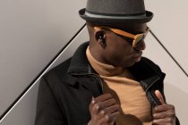 Moda confiante afro-americano masculino em chapéu e óculos de sol em pé contra a parede cinza e olhando para baixo — Fotografia de Stock