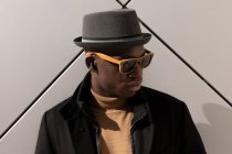 Trendy selbstbewusster afroamerikanischer Mann mit Hut und Sonnenbrille steht vor grauer Wand und blickt nach unten — Stockfoto