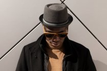 Moda confiante afro-americano masculino em chapéu e óculos de sol em pé contra a parede cinza e olhando para baixo — Fotografia de Stock
