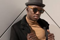 Moda confiante afro-americano masculino em chapéu e óculos de sol em pé contra a parede cinza e olhando para a câmera — Fotografia de Stock