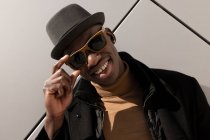 Moda confiante feliz afro-americano masculino em chapéu e óculos de sol em pé contra a parede cinza e olhando para a câmera — Fotografia de Stock