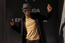 Trendy confident happy African American male in hat and sunglasses standing and dancing against gray wall and looking at camera — Stock Photo