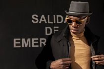 Впевнений афроамериканець у капелюсі й сонцезахисних окулярах стоїть проти сірої стіни й озирається геть. — стокове фото