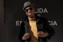 Trendy selbstbewusster afroamerikanischer Mann mit Hut und Sonnenbrille steht vor grauer Wand und blickt in die Kamera — Stockfoto
