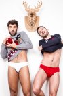 Couple of adult gay lovers in underwear and Xmas sweaters with decorative red balls standing against white wall with artificial deer head — стоковое фото