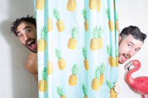 Веселые взрослые бородатые гомосексуальные бойфренды с мокрыми волосами и мыльной пеной, смотрящие в занавеску, принимая душ вместе — стоковое фото