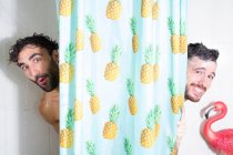 Веселые взрослые бородатые гомосексуальные бойфренды с мокрыми волосами и мыльной пеной, смотрящие в занавеску, принимая душ вместе — стоковое фото