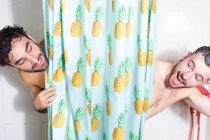 Alegre adulto barbudo homosexual novios con el pelo mojado y espuma de jabón mirando hacia fuera cortina mientras toma ducha juntos - foto de stock