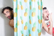 Cheerful adult bearded homosexual boyfriends with wet hair and soap foam looking out curtain while taking shower together — Stock Photo