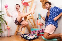 Lustig aufgeregt diverse erwachsene homosexuelle Freunde in sommerlichen Outfits mit Drinks, die vorgeben, mit rosa Flamingo am Strand zu sein und zusammen Spaß zu haben — Stockfoto
