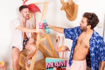 Funny excited diverse adult homosexual boyfriends in summer outfits with drinks pretending being on beach with pink flamingo and having fun together — Stock Photo