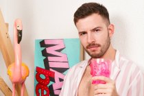Молодий бородатий гомосексуальний хлопець в літньому вбранні п'є коктейль з рожевої чаші, сидячи на підлозі вдома і прикидаючись на пляжі — стокове фото