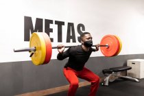 Determined young African American male athlete in activewear and face mask lifting heavy metal barbell while training in gym during coronavirus pandemic — Stock Photo