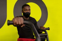 Jovem atleta masculino afro-americano concentrado em sportswear e máscara facial exercitando-se na máquina de ciclismo durante o treinamento em ginásio contra fundo amarelo brilhante — Fotografia de Stock
