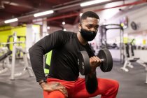 Joven deportista afroamericano confiado en ropa deportiva y máscara levantando pesadas mancuernas mientras está sentado en el banco en el gimnasio moderno - foto de stock