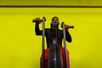 Konzentrierter junger afroamerikanischer Athlet in Sportbekleidung und Gesichtsmaske, der während des Trainings im Fitnessstudio vor leuchtend gelbem Hintergrund auf einem Fahrrad trainiert — Stockfoto