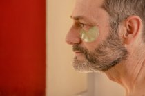 Side view of cropped bearded middle aged male with naked torso applying eye patch on skin while looking in mirror at home — Stock Photo