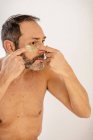 Cultiver mâle d'âge moyen barbu avec torse nu appliquant un bandeau oculaire sur la peau tout en regardant dans le miroir à la maison — Photo de stock