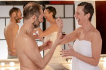 Sid view of cheerful female in towel applying black peel off mask on face of male beloved while looking at each other reflecting in mirror at home — Stock Photo