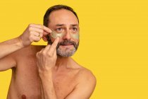 Homem de meia idade com tronco nu aplicando manchas oculares na pele enquanto olha para o fundo amarelo — Fotografia de Stock