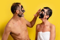 Alegre macho en toalla aplicando la máscara de piel negra en la cara de la mujer amada mientras se miran el uno al otro en el fondo amarillo - foto de stock
