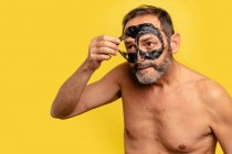 Чоловік середнього віку з голим торсом чистить чорну маску від обличчя, дивлячись на жовтий фон — стокове фото