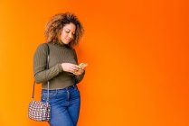 Retrato de una mujer afro usando móvil con una pared naranja en el fondo - foto de stock