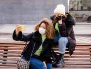Дві жінки друзі беруть автопортрет з мобільним телефоном, одягнені в захисну маску на вулиці — стокове фото