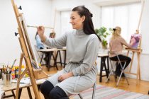 Side view of focused female artist painting on canvas on easel in art studio on background of blurred women — Stock Photo