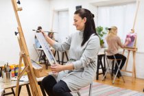 Vue latérale de la peinture d'artiste féminine concentrée sur toile sur chevalet dans un atelier d'art sur fond de femmes floues — Photo de stock