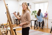 Side view of delighted female artist looking at camera while painting on canvas on easel in art studio on background of blurred women — Stock Photo
