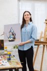 Entzückte Künstlerin steht mit Malerei auf Leinwand in Kreativwerkstatt und blickt in die Kamera — Stockfoto