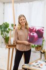 Artista femenina encantada de pie con pintura sobre lienzo en taller creativo y mirando a la cámara - foto de stock