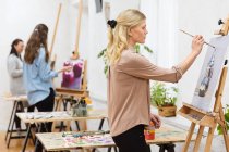 Vista laterale della pittura focalizzata artista femminile su tela su cavalletto in studio d'arte su sfondo di donne sfocate — Foto stock