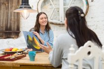 Cheerful female artists drinking beverages while discussing painting at table in art workshop — Stock Photo