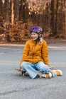Souriant adolescent fille en casque assis sur skateboard sur le parking tout en profitant week-end en automne et en regardant loin — Photo de stock
