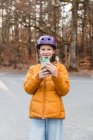 Веселая девушка-подросток в шлеме фотографирует на мобильный телефон, стоя на парковке в осеннем парке и глядя в камеру — стоковое фото