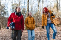Coppia sorridente e figlia adolescente che cammina con cesto e plaid nel bosco per fare un picnic in autunno — Foto stock