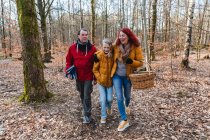 Coppia sorridente e figlia adolescente che cammina con cesto e plaid nel bosco per fare un picnic in autunno — Foto stock