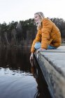 Low angle of cheerful teenage girl sitting on wooden quay near pond in autumn forest and looking away — Stock Photo