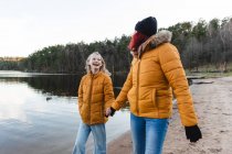 Zufriedene Mutter und Teenager halten Händchen und spazieren am Teich im Wald entlang, während sie das Herbstwochenende genießen — Stockfoto