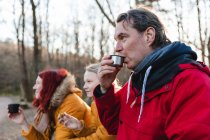 Vue latérale de la famille heureuse buvant du thé dans des tasses thermos et mangeant de la nourriture tout en profitant du pique-nique dans les bois en automne — Photo de stock