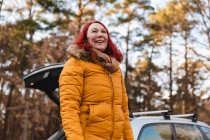 Low angle of cheerful female traveler in outerwear standing near car woods and looking away — Stock Photo
